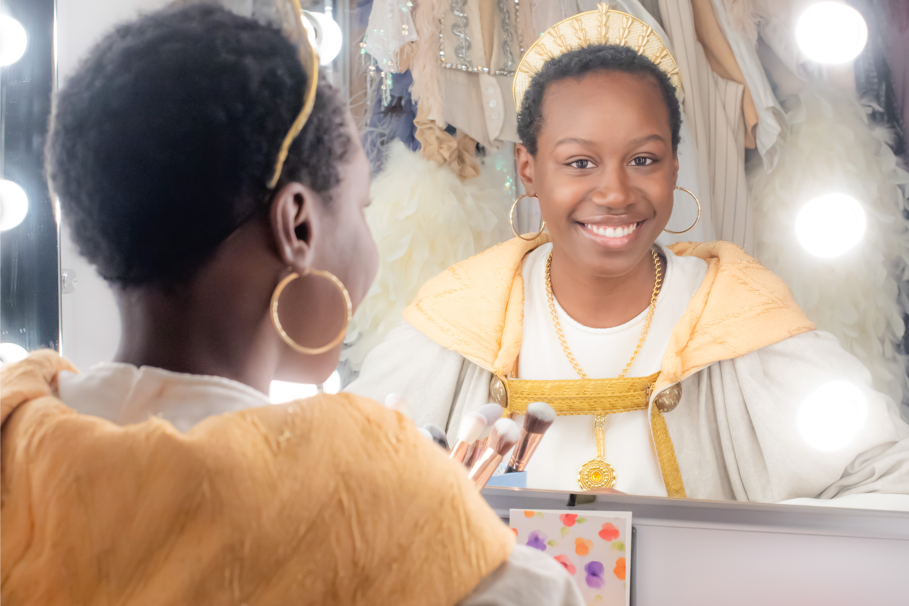 A black women wearing a costume, with a gold headpiece, smiling at herself in the mirror in the dressing rooms.